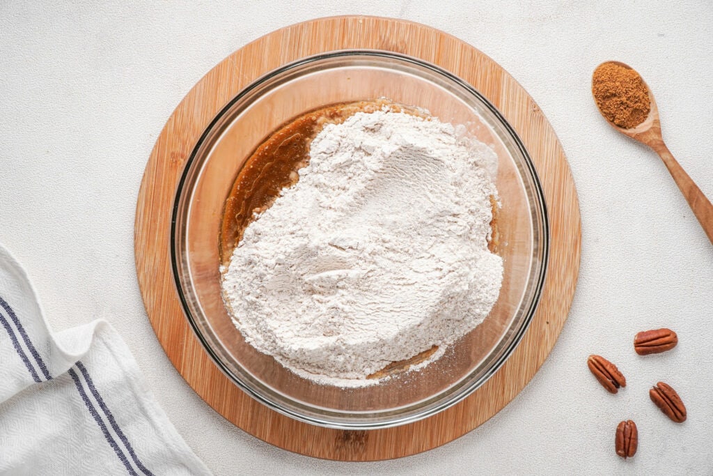 Wet and dry ingredients in a glass bowl on a wooden round board with four pecans and a wooden spoon with pumpkin pie spice to the right.