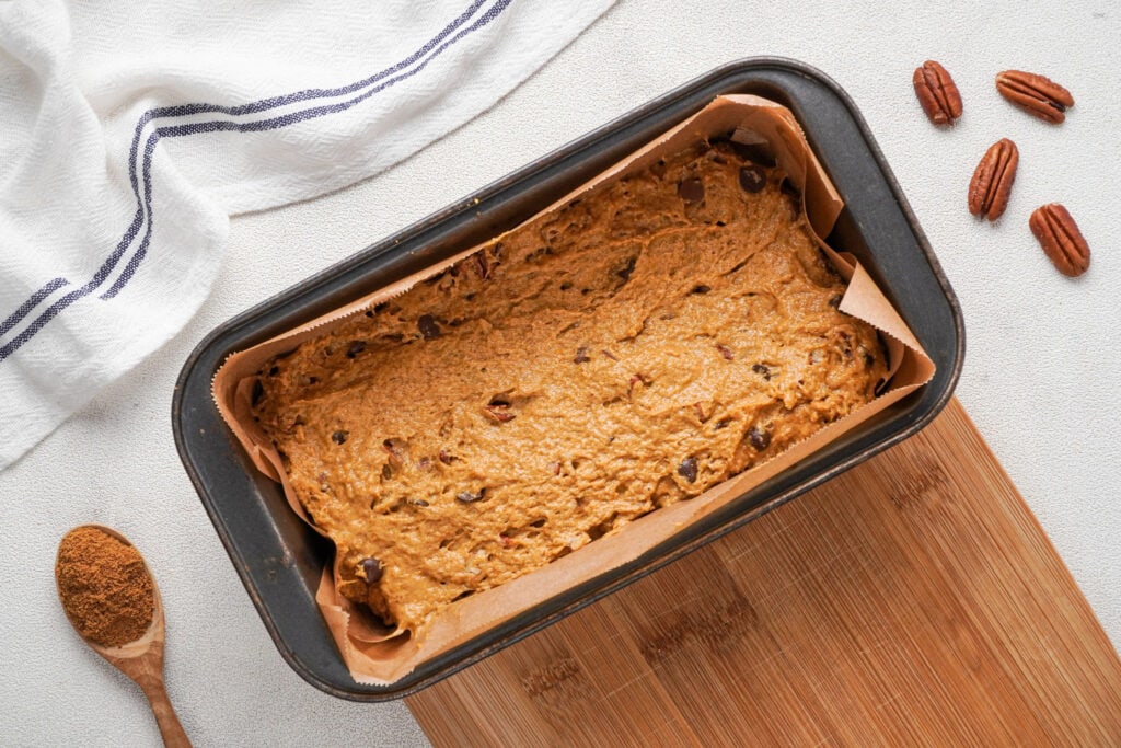 A loaf pan on a bread board with the pumpkin bread batater ready to bake with a blue striped cloth to the top, a wooden spoon with spices at the bottom left corner and four pecan halves on the top right corner.