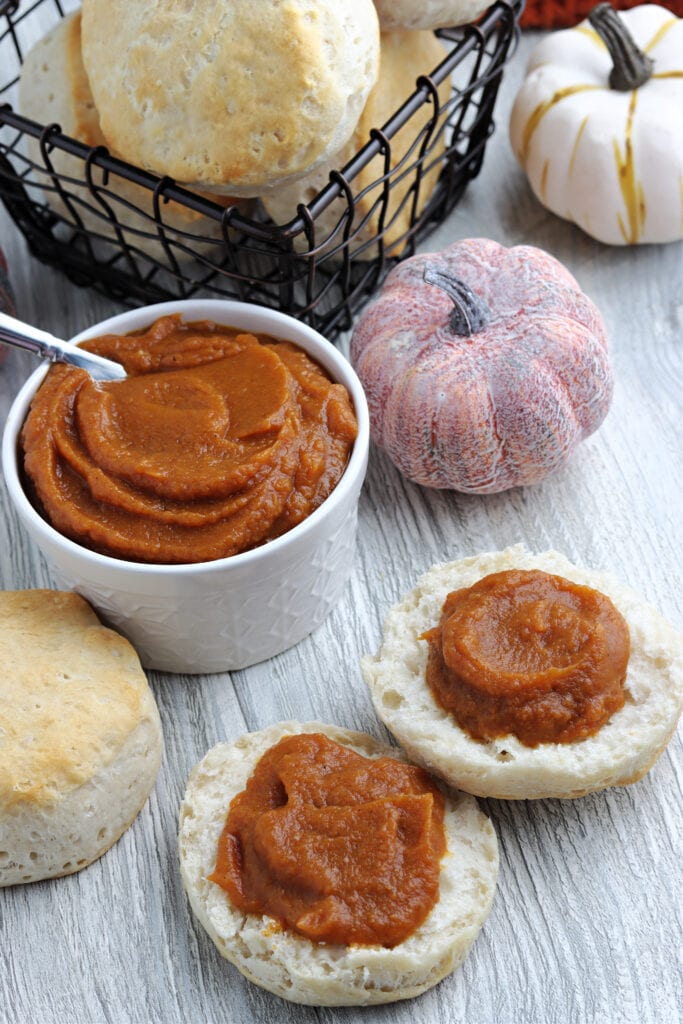 White bowl with pumpkin butter. Two biscuit halves with a generous helping of pumpkin butter spread on each with two ceramic pumpkin props to the right and a black wire basket of biscuits to the back.