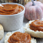 White bowl with pumpkin butter. Two biscuit halves with a generous helping of pumpkin butter spread on each with a ceramic pumpkin prop to the right.