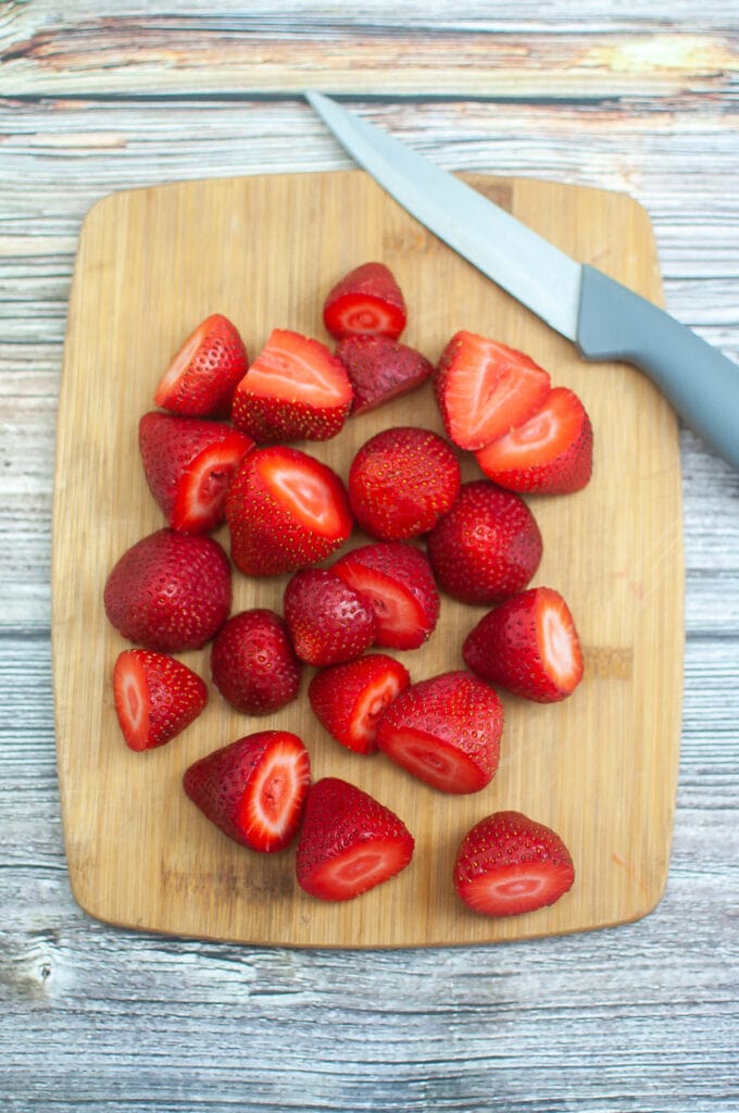 Cut hulled strawberries on a wooden cutting board. A knife to the top right corner.