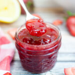Strawberry jam being spooned from a glass jar with strawberries, lemon half, and a red checked cloth surrounding.