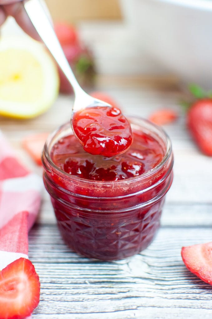 Strawberry jam being spooned from a glass jar with strawberries, lemon half, and a red checked cloth surrounding.