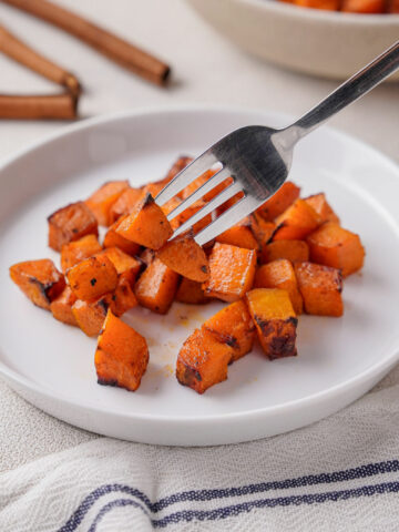 A white bowl of air fried butternut squash in right upper corner with a smaller white plate of butternut squash in right lower corner with cinnamon sticks surrounding.