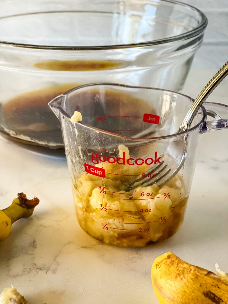 Mashing the banana into the measuring cup with a fork. Bananas in the foreground and a glass bowl with liquid ingredients in the background.