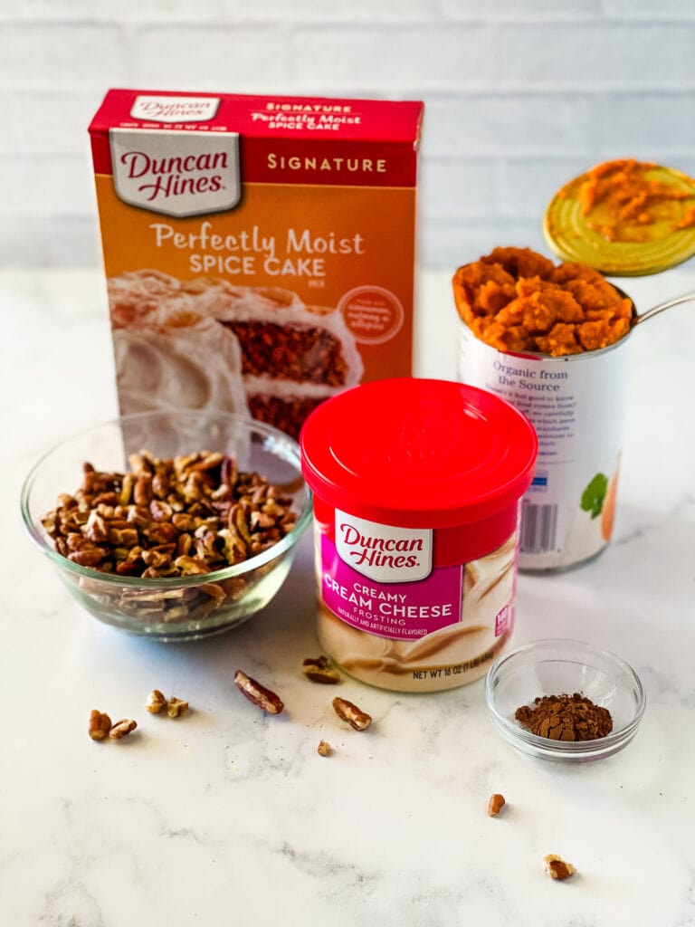 The ingredients needed to make a pumpkin cake including a cake mix, prepared frosting, chopped pecans, pumpkin puree and pumpkin pie spice. A few pecans are scattered around.