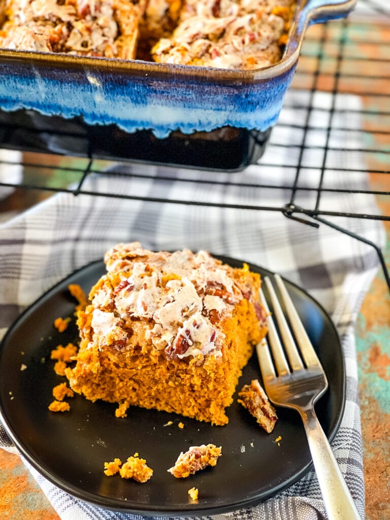 A slice of vegan pumpkin cake on a black plate with a cup of coffee, pumpkins, and a gray checked cloth in the foreground and background.