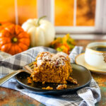 A slice of vegan pumpkin cake on a black plate with a cup of coffee, pumpkins, and a gray checked cloth in the foreground and background.