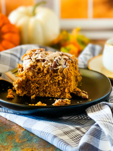 A slice of vegan pumpkin cake on a black plate with a cup of coffee, pumpkins, and a gray checked cloth in the foreground and background.