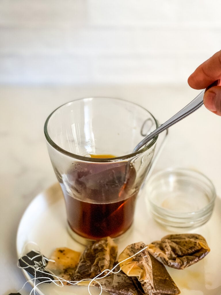 Brewed tea in a glass cup with a hand holding a spoon in it with used tea bags on the plate.