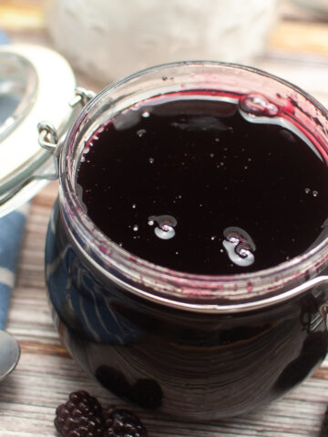 An open jar of blackberry jam with a blue striped cloth to the left and blackberries on the counter in front of the jar on the counter.