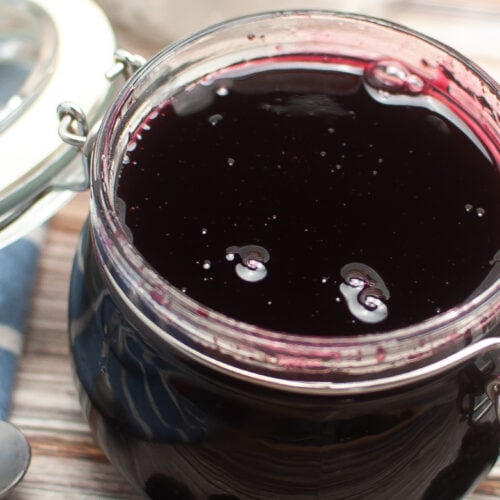 An open jar of blackberry jam with a blue striped cloth to the left and blackberries on the counter in front of the jar on the counter.