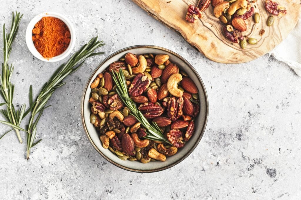 Bowl of mixed roasted nuts with spring of rosemary. Rosemary and spices on the left side. A wooded cutting board with mixed nuts.