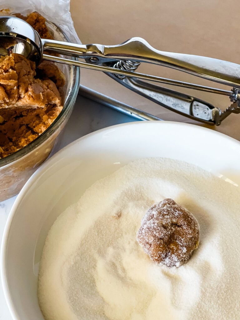 A white bowl of sugar with one dough ball in the sugar having been rolled. A glass bowl with dough and a cookie scoop in the rear left corner.