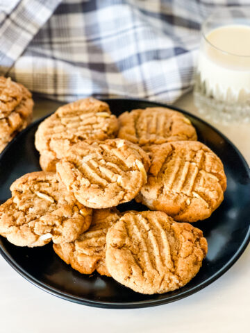 A black plate with several almond butter cookies with a gray checked cloth and a glass of milk in the back.