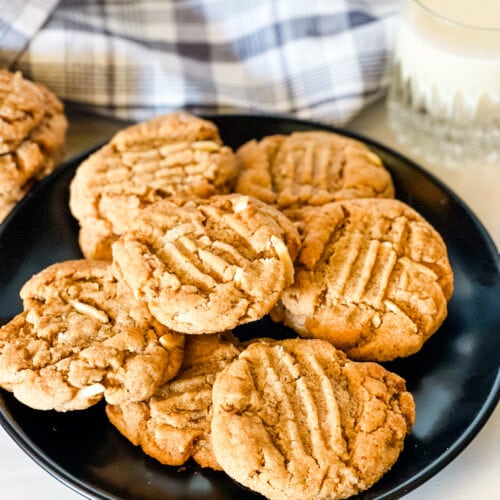 A black plate with several almond butter cookies with a gray checked cloth and a glass of milk in the back.