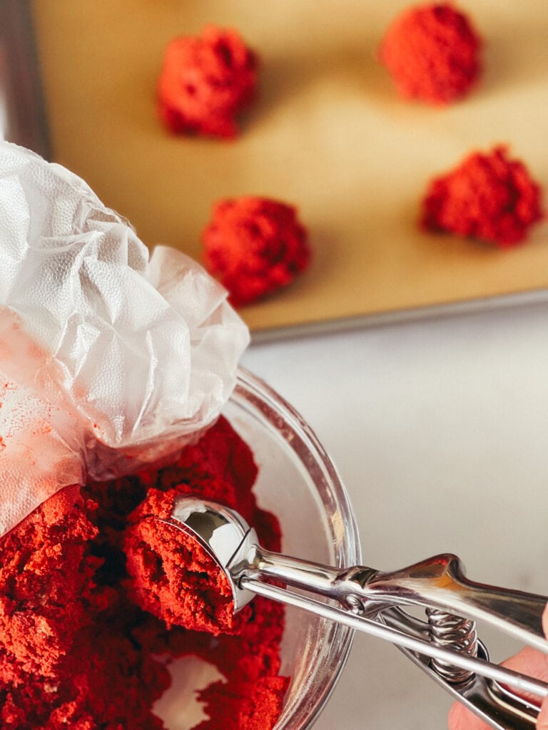 Removing a ball of red vevet cookie dough from the bowl of raw dough with a cookie scoop. Some are on the cookie sheet behind it.