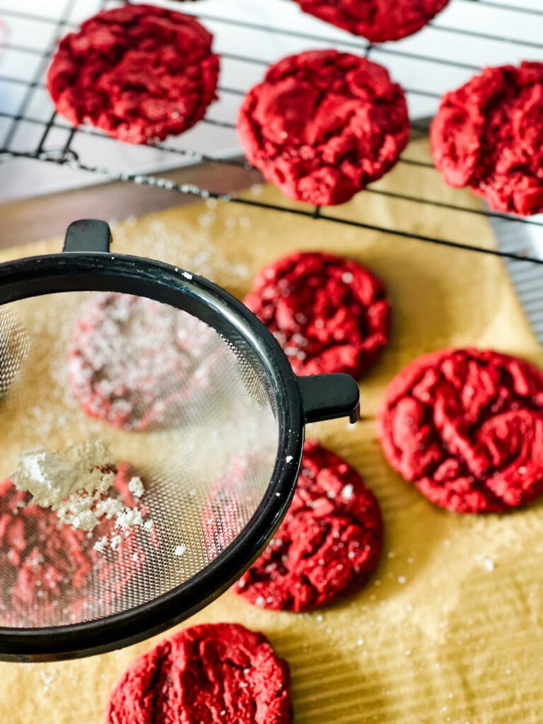 Sprinkling the powdered sugar on the red velvet cookies with a mesh strainer. Some cookies are on the baking sheet and some cookies are on the wire rack.