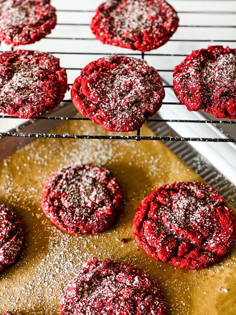 A cookie sheet with some red velvet cookies and a cooling rack above it with some red velvet cookies. All covered with powdered sugar.