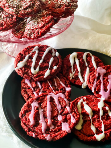 A pink raised plate with vegan red velvelt cookies on a white cloth with more cookies on a black plate. The cookies on the glass plate have powdered sugar topping and the cookies on the black plate have drizzles of icing.
