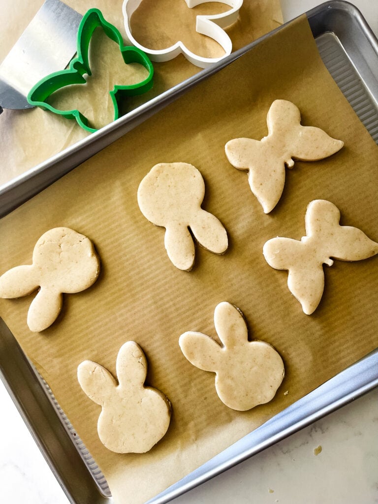 Four bunny and two butterfly coolies on a parchment paper covered cookie sheet ready to bake. The two cookie cutters are at the top.