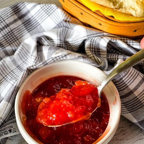 Spooning out some strawberry pineapple jam from a white dish with a basket of rolls in the background.