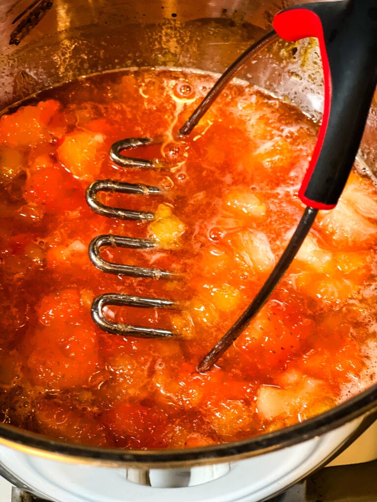 Mashing the cooked ingredients for strawberry pineapple jam with a potato masher.
