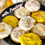 Plate of vegan lemon sugar cookies - some with white frosting and some with yellow and all with sprinkles on a black plate with a glass of plant milk in the background with Pinterest text overlay.