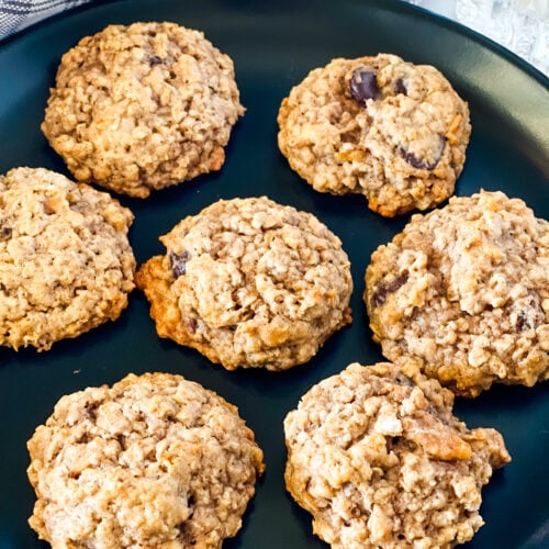Vegan Banana Oatmeal Cookies on a black plate with a gray checked napkin and glass of plant milk in the rear.