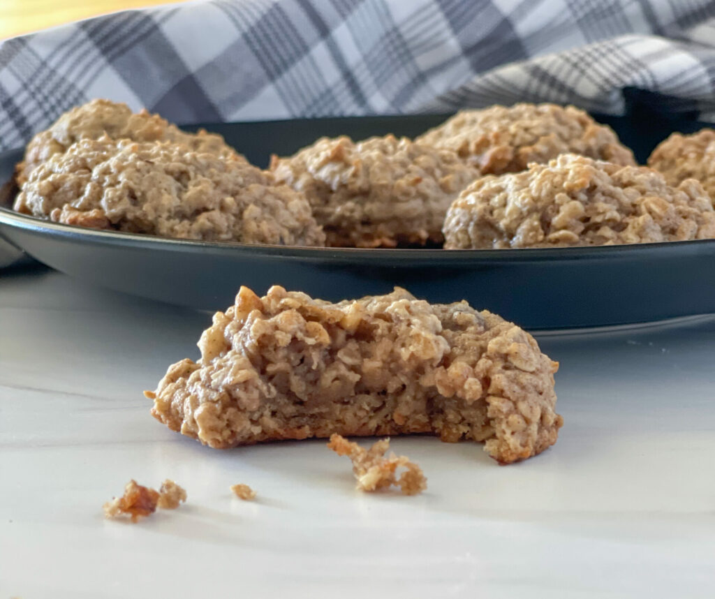 Vegan Banana Oatmeal Cookies on a black plate with a gray checked napkin in the rear. One cookie is on the counter and has a bite taken out from it.