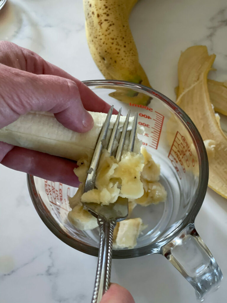 Mashing and adding the banana with a fork into a glass measuring cup. Banana peels are in the background.