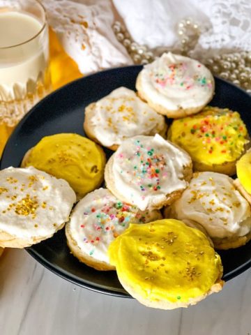 Plate of vegan lemon sugar cookies - some with white frosting and some with yellow and all with sprinkles on a black plate with a glass of plant milk in the background.
