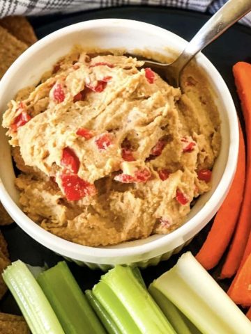 White bowl with vegan pimento cheese spread with a spoon dipping in and carrot and celery sticks at the bottom and side with a gray checked cloth in the background.