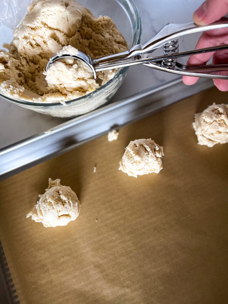 Using a cookie scoop to scoop out a dough ball of lemon cookie dough. A parchment paper lined cookie sheet with three cookie dough balls is also in the photo.