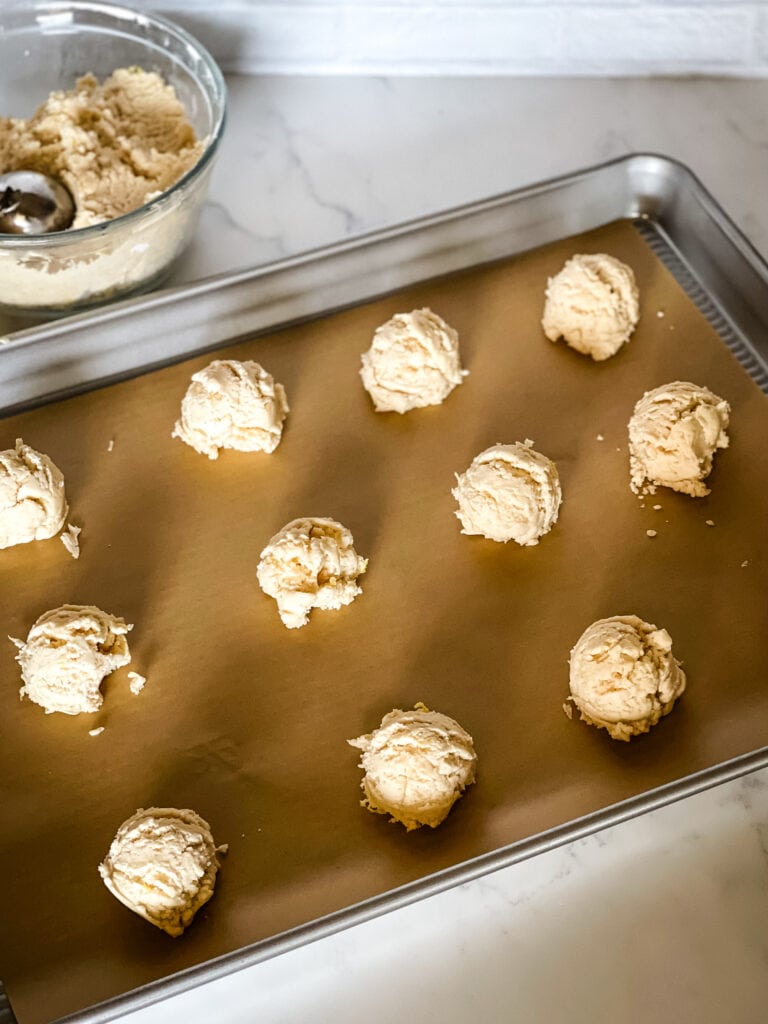 A parchment paper lined cookie sheet with twelve lemon sugar cookie dough balls and a bowl of unbaked dough with a cookie scoop in the top left corner.