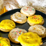 Plate of vegan lemon sugar cookies - some with white frosting and some with yellow and all with sprinkles on a black plate with a glass of vegan milk in the background.