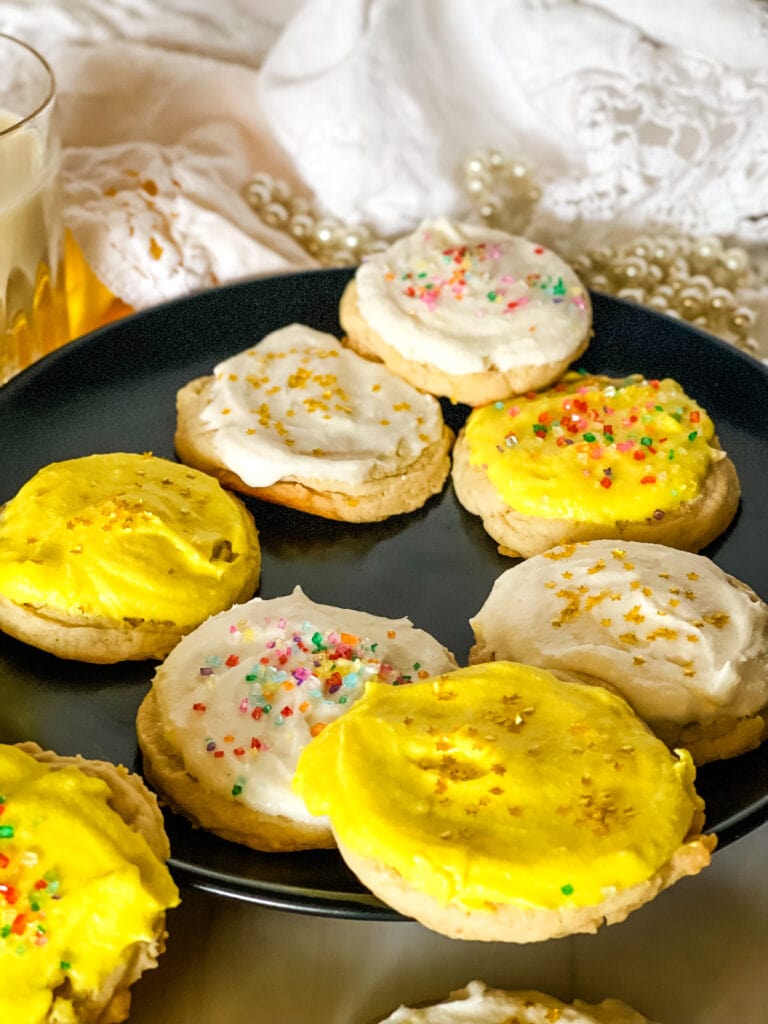 Plate of vegan lemon sugar cookies - some with white frosting and some with yellow and all with sprinkles on a black plate with a glass of vegan milk in the background.