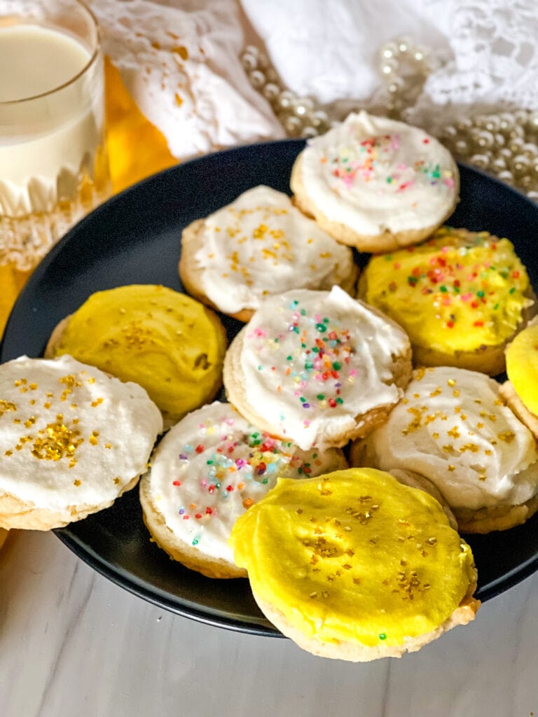 Plate of vegan lemon sugar cookies - some with white frosting and some with yellow and all with sprinkles on a black plate with a glass of plant milk in the background.