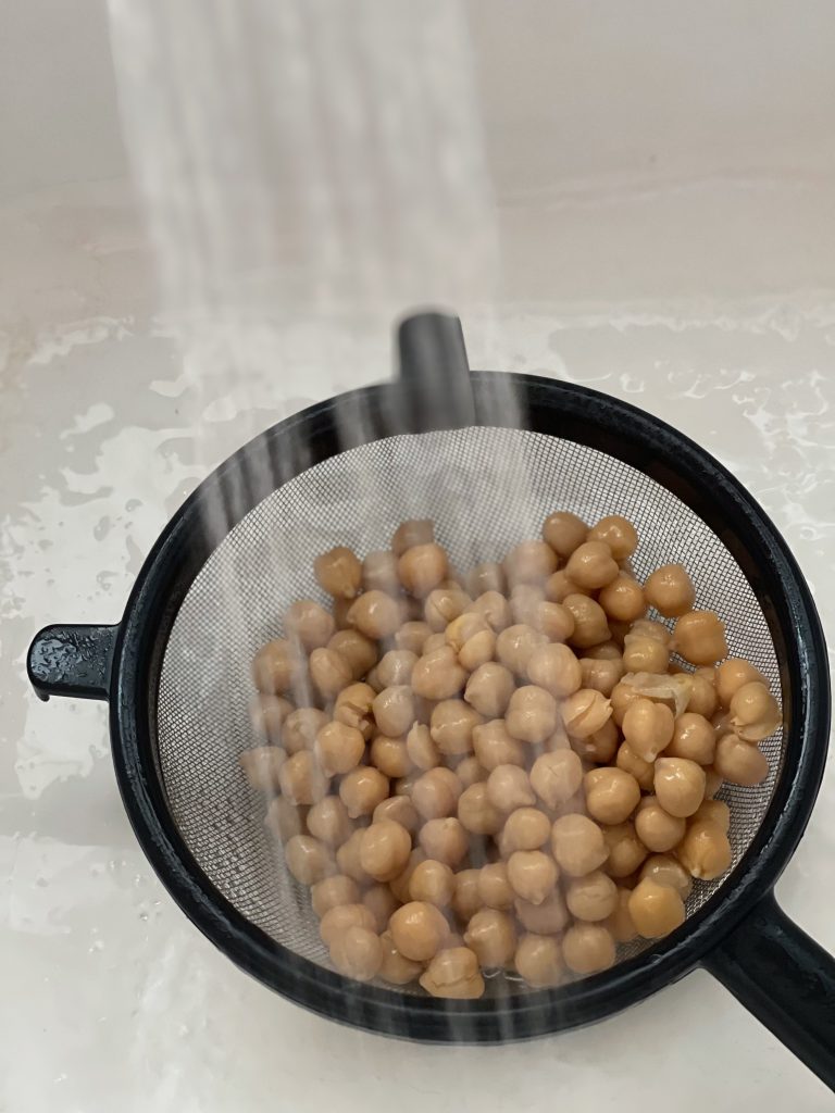 Rinsing the chickpeas in the sink with a stream of water over the wire mesh sieve containing canned chickpeas.