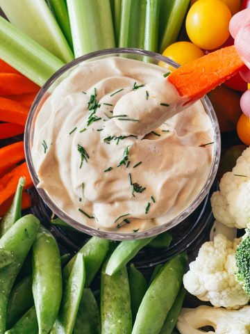 A bowl of tofu dip with a carrot dipping out a portion surrounded by various veggies.
