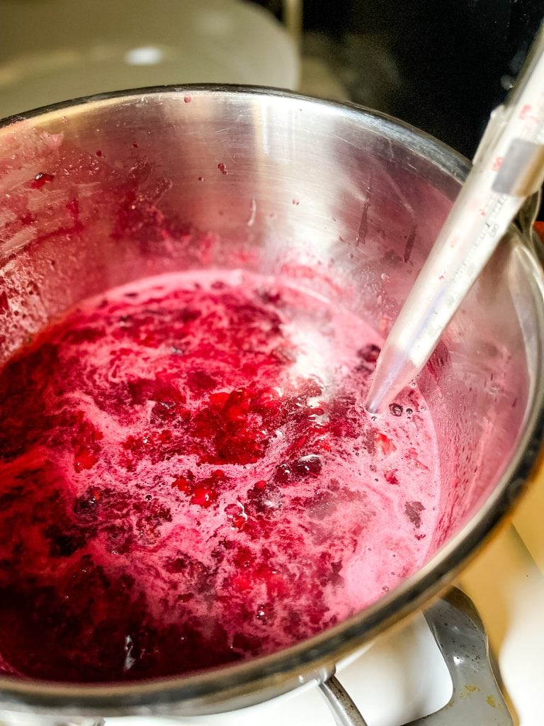 Showing the triple berry jam boiling in the pan with a candy thermometer on the side.