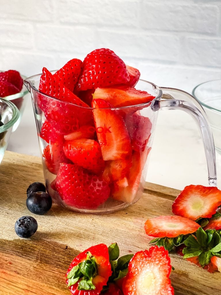 A generous cup of cut strawberries next to some other glass bowls with a few blueberries on the wooden cutting board and cut tops of strawberries.