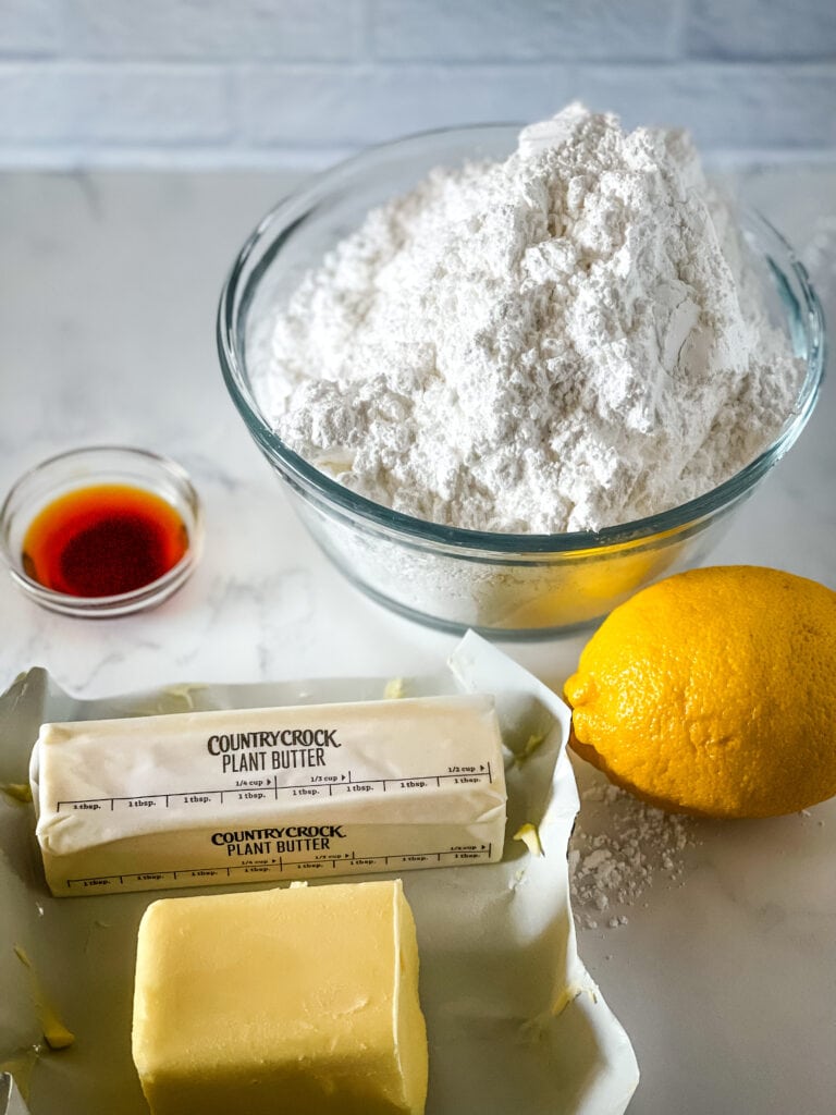Ingredients needed for vegan lemon frosting including vanilla extract, vegan butter, butter flavored shortening, a lemon and powdered sugar.