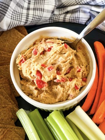 White bowl with vegan pimento cheese spread with a spoon dipping in and carrot and celery sticks at the bottom and side with a gray checked cloth in the background.