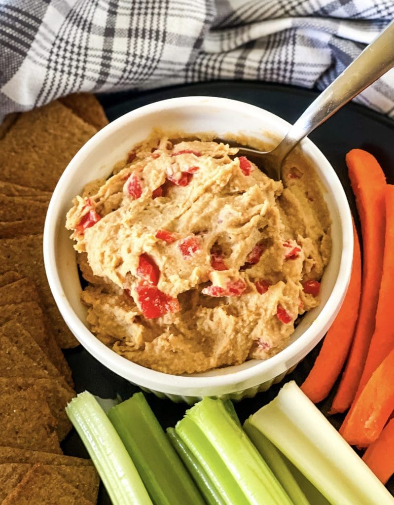 White bowl with vegan pimento cheese spread with a spoon dipping in and carrot and celery sticks at the bottom and side with a gray checked cloth in the background.