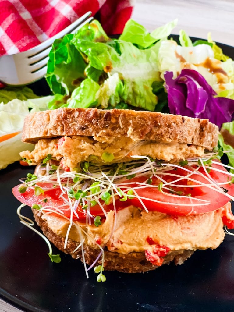 Vegan pimento cheese spread on two slices of whole wheat toast with tomato and sprouts with salad, fork, and red checked napkin in the background.