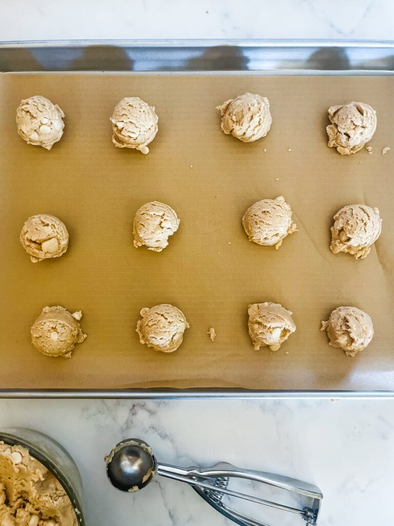 A parchment paper lined cookie sheet with twelve uncooked white chocolate chip macadamia nut dough balls on it with a bowl of dough and a cookie scoop in front.