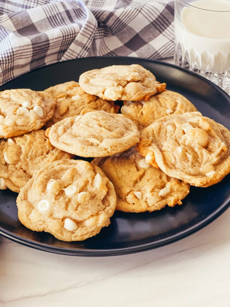 Black plate with vegan white chocolate macadamia nut cookies with gray checked cloth and glass of oat milk in the rear.