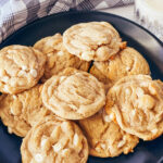 Black plate with vegan white chocolate macadamia nut cookies with gray checked cloth and glass of oat milk in the rear.