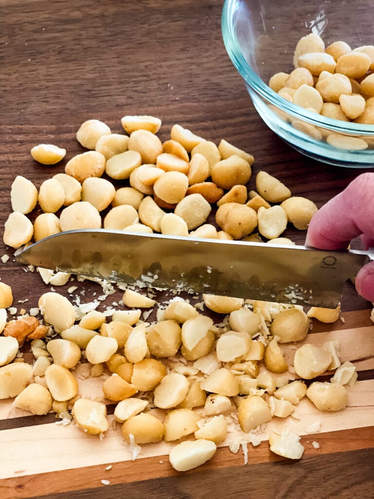 Chopping the macadamia nuts with a knife on a cutting board with a bowl of macadamia nuts top right.